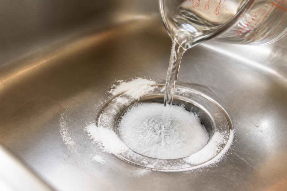 Wlewanie wody do odpływu zlewu Miarka z wodą nad odpływem zlewu otoczonym białym proszkiemWe need answer in Polish exact format. Need unique. Image pouring water into drain with powder likely soda but existing says don't repeat. Need maybe "Przepłukiwanie odpływu zlewu wodą". Alt concise. Ensure not mention soda? unique from existing. Let's answer.